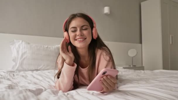 A pretty nice young girl is listening to music using her smartphone and earphones lying in the bed and at home - Felvétel, videó