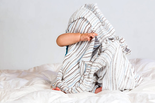 Small Caucasian boy playing under a striped blanket on white duvet bedding - 写真・画像