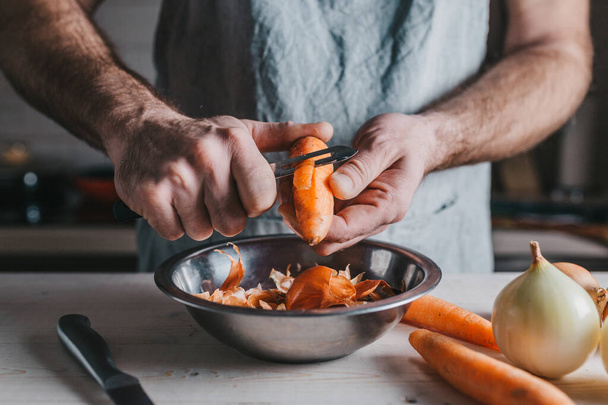 Primer plano chef profesional con las manos ásperas limpia una zanahoria naranja madura - sobre el fondo de una cocina rústica
 - Foto, Imagen
