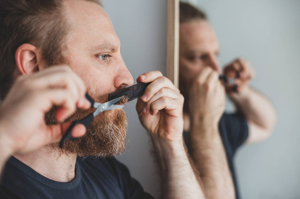 隔離中のセルフケア。大人のハンサムな白人男性が口ひげを切り、自宅ではさみで自分自身をひげを生やしています。コロナウイルスの発生と人々のシャットダウン、家庭の概念を維持する - 写真・画像