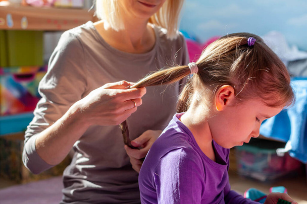 Mother styling her little daughters hair, holding it doing her ponytails. Mom and daughter close intimate moments. Children, kids hair, sunlight, kids room, two people lifestyle, family moment - Photo, Image