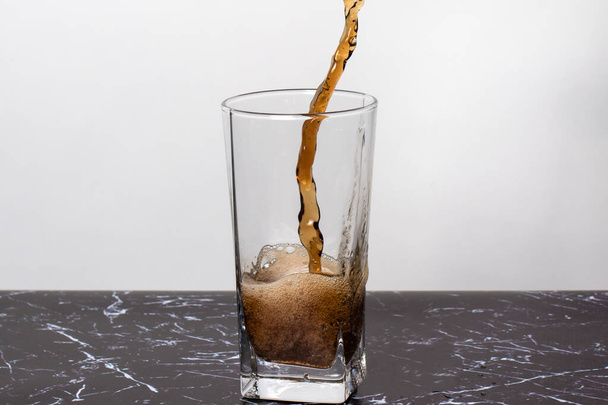 Ένα καφέ σκούρο γλυκό ποτό χύνεται σε ένα ποτήρι ποτήρι ζέσεως, πιτσιλιές και μια βουτιά ροφήματος. - Φωτογραφία, εικόνα