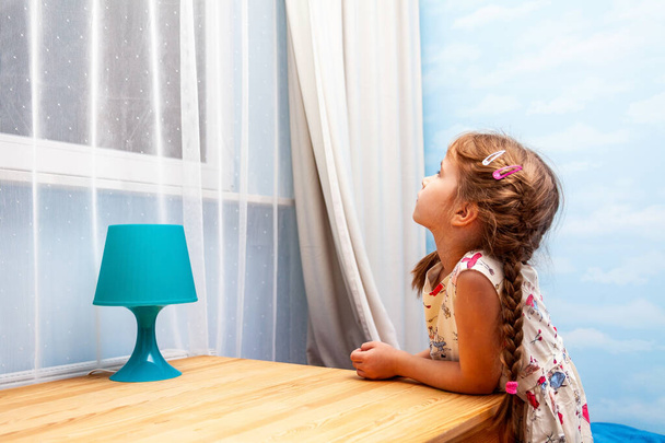 Маленькая девочка, ребенок, прислонившись к столу, смотрит в окно ночью в одиночестве в светлой комнате. Ждать, когда кто-то вернется, искать, мечтать, надеяться.
 - Фото, изображение