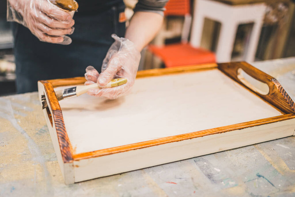 Carpentry workshop - master applies varnish and tints wooden craft serving tray - 写真・画像