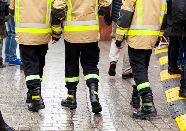 Drie anonieme brandweerman in gele reflecterende uniformen en zwarte laarzen en broeken lopen weg van de camera op de drukke straat, achterkant, benen. Generieke geüniformeerde diensten - Foto, afbeelding