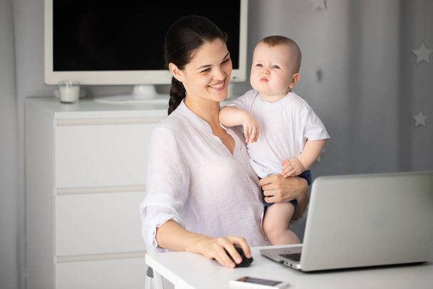 Young mother working in a decree at the computer with a baby son in her arms - Φωτογραφία, εικόνα
