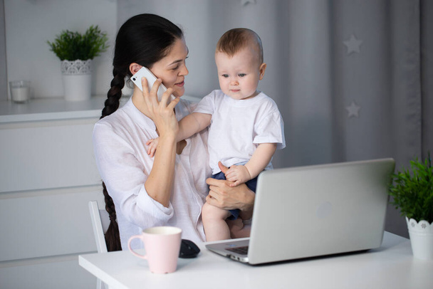 Nuori äiti työskentelee asetuksessa tietokoneella vauva poika sylissään
 - Valokuva, kuva