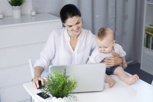 Fiatal anya egy rendeletben dolgozik a számítógépnél egy kisfiúval a karjaiban. - Fotó, kép