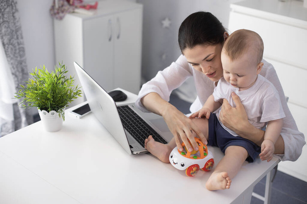 Fiatal anya egy rendeletben dolgozik a számítógépnél egy kisfiúval a karjaiban. - Fotó, kép
