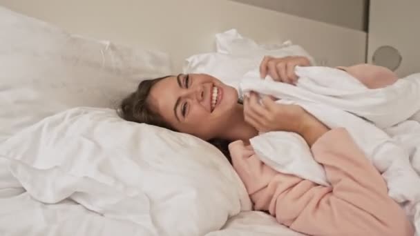 A smiling young girl is having fun and laughing laying in the bed at home - Filmati, video