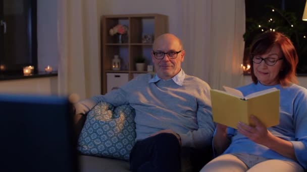 senior couple watching tv and reading book at home - Materiał filmowy, wideo