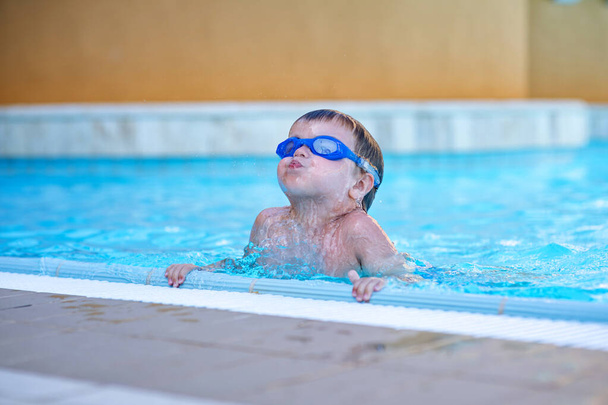 Lapsi, jolla on uimalasit, nousee vedestä. Sinisilmäinen poika pitää hauskaa altaassa. Kesäulkoilu. Uinti altaassa kirkkaalla sinisellä vedellä
. - Valokuva, kuva