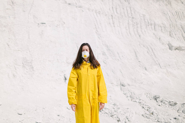 Young woman walking on a phosphate mountain that was made by chemical plant waste. She is wearing yellow protective coat and a mask. Ecology protection awareness - Zdjęcie, obraz