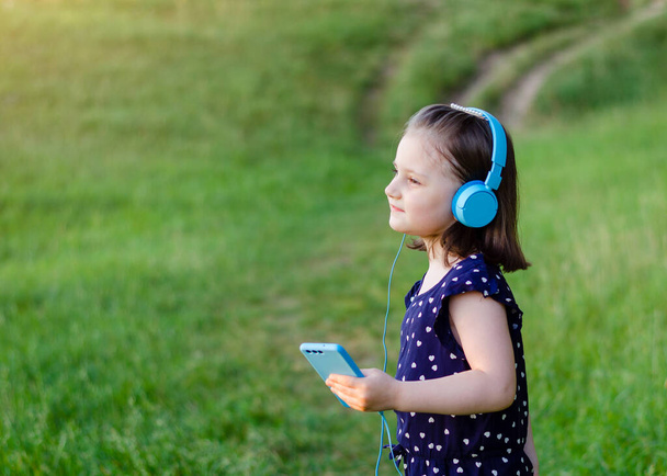 A little girl in blue headphones with a smartphone in her hands is dancing on the background of a field and green grass in the summer at sunset. Girl dressed in a blue jumpsuit with hearts - Photo, Image