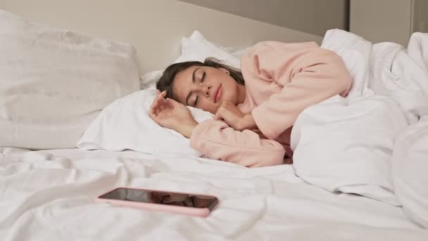 A young sleepy girl is turning off her alarm clock on her mobile phone laying in the bed at home - Video