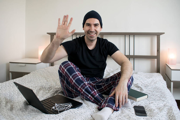 jovem macho sentar na cama criador de conteúdo doméstico smartphone laptop mão onda sorriso fones de ouvido e livro
 - Foto, Imagem