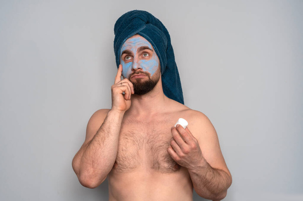Бородатый мужчина с синим полотенцем на голове и косметической маской на лице. Косметологические процедуры для мужчин. Уход за кожей для мужчин
. - Фото, изображение
