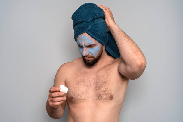 Um homem barbudo com uma toalha azul na cabeça e uma máscara cosmética no rosto. Procedimentos de cosmetologia para homens. Cuidados com a pele para homens
. - Foto, Imagem