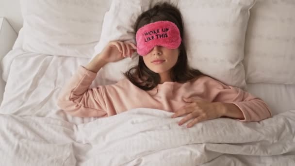 A lovely cheerful girl is taking off her sleeping mask laying in the bed at home - Filmati, video