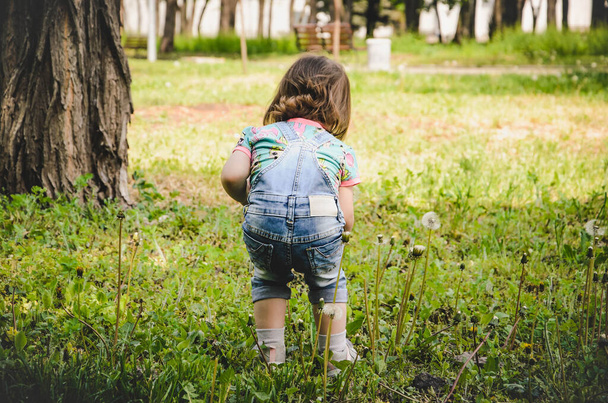 květen 2020. Kyjevská Ukrajina. Malá holčička si hraje venku v parku, sbírá rostliny za slunečného dne v džínových montérkách a tričku s potiskem LOL - Fotografie, Obrázek