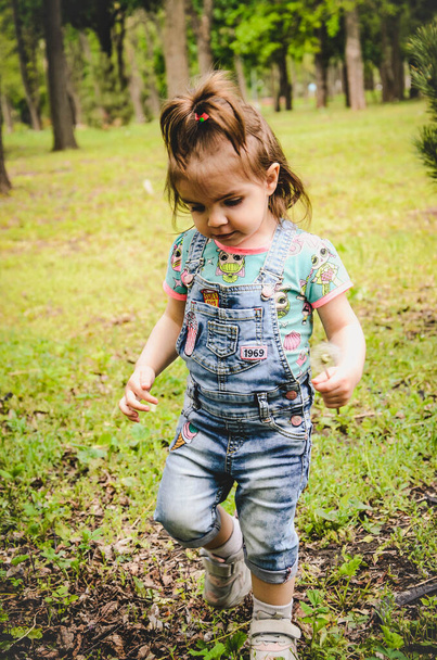 May 2020. Kiev Ukraine. A little girl plays outside in a park, collects plants on a sunny day in denim overalls and a T-shirt with a print LOL - Foto, imagen