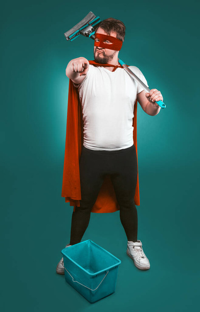 Superhero is ready to fight for cleanliness, Brave man in a red superhero uniform with a bucket and a mop points the camera with his index finger. Cleaning concept. Isolated on green - Foto, immagini