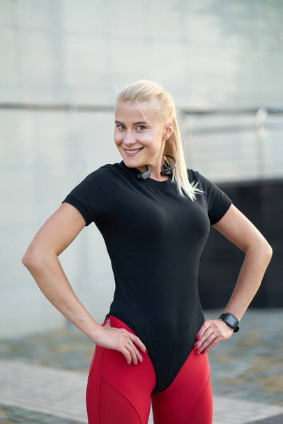 Portrét mladé atraktivní blondýny pozitivní dívka v černém sportovním oblečení a módní červené kamaše. Venkovní záběr na šedé pozadí zdi.                               - Fotografie, Obrázek