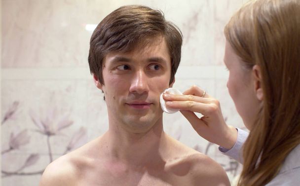 Close up portrait of young handsome man whose wife wipes his face with a cotton pad in the bright bathroom. Male face skin care. Man making funny faces - Foto, afbeelding