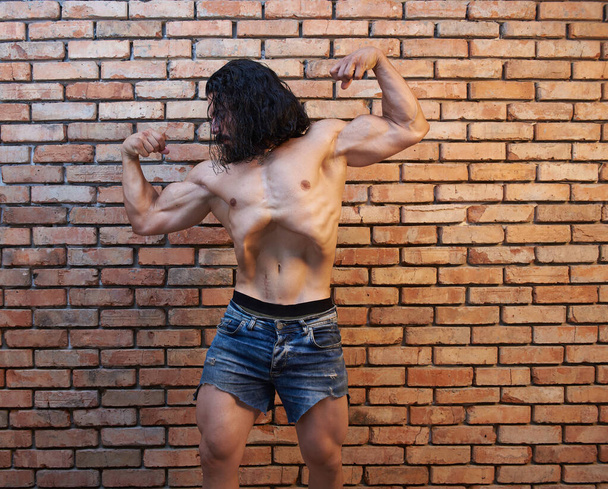 jovem homem musculoso em jeans curtos posando contra a parede de tijolo, homem jovem muscular, homem jovem muscular posando
 - Foto, Imagem