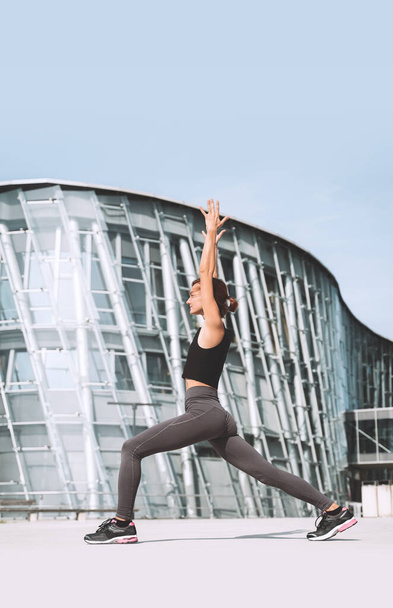 Beautiful woman doing stretching yoga exercises outside in the city. Young girl practicing yoga outdoors on background of modern skyscrapers. Concept of healthy lifestyle, sport, urban life. - Foto, imagen