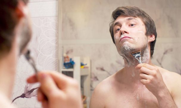 Close-up face of handsome young man shaving his face. Portrait of man reflected in the mirror the in bathroom. Daily face care - Foto, afbeelding