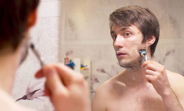 Een close-up van een knappe jongeman die zijn gezicht scheert. Portret van de mens weerspiegeld in de spiegel de in badkamer. Dagelijkse gezichtsverzorging - Foto, afbeelding