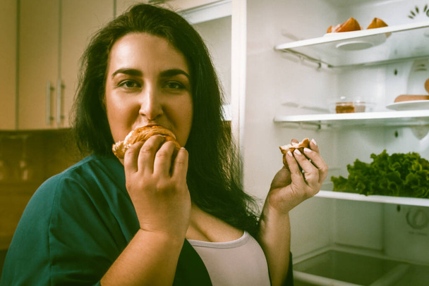 Mujer con sobrepeso come comida poco saludable, mujer caucásica hambrienta come o devora un croissant mirando a la cámara
 - Foto, Imagen