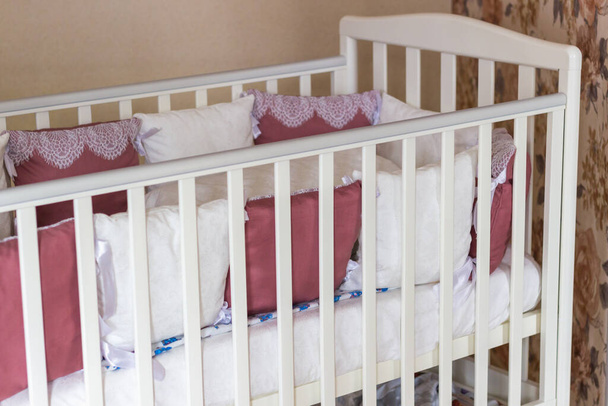 Babybett mit weißen und bordeauxfarbenen Kissen mit Schnürsenkeln. Kinderbett auf Rädern im Schlafzimmer mit pastellgelben Farben. Minimalistisches Interieur - Foto, Bild