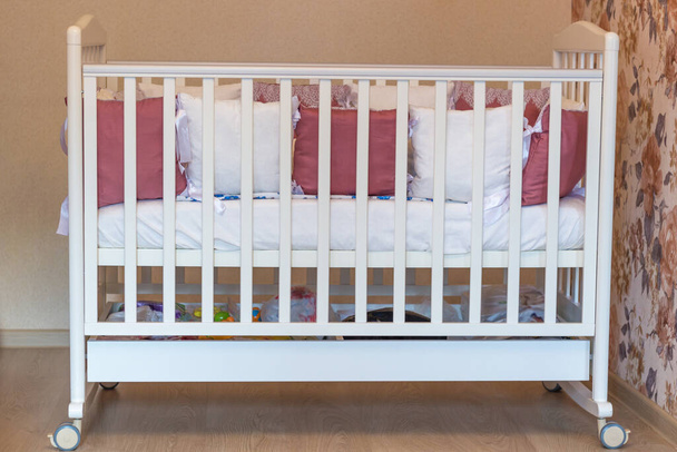 Cama de bebé con almohadas de color blanco y borgoña con cordones. Cuna sobre ruedas en el dormitorio con colores amarillos pastel. Interior minimalista
 - Foto, Imagen
