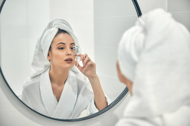 Beautiful young woman using curlier for eyelashes in bathroom - Фото, изображение