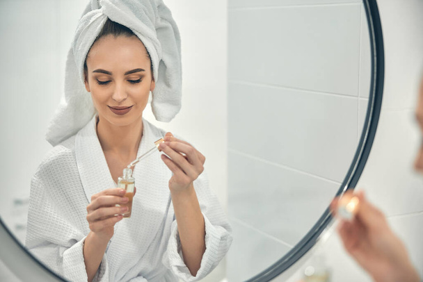 Caucasian woman with white towel on head using face serum in bathroom - Fotó, kép