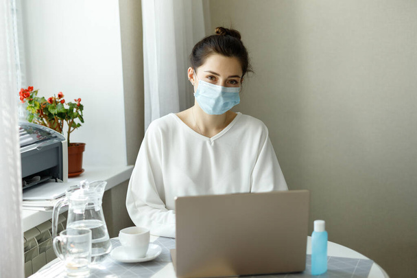 young woman girl student in protective medical mask, hand sanitizer on the table, working with a laptop notebook, online learning education shopping, work at home during quarantine covid-19 - Photo, image