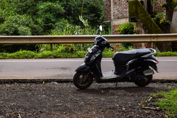 Фотографії чорного кольору скуті або скутер припарковані на стороні дороги. порожній асфальт і зелені дерева на задньому плані. Знімок виконано в Колхапурі (Махараштра).. - Фото, зображення