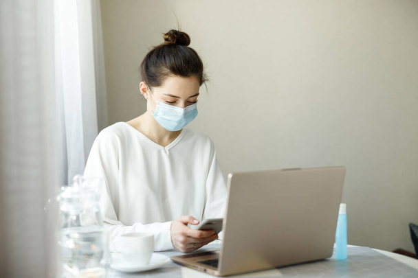 young woman girl student in protective medical mask, hand sanitizer on the table, working with a laptop notebook, online learning education shopping, work at home during quarantine covid-19 - Photo, Image