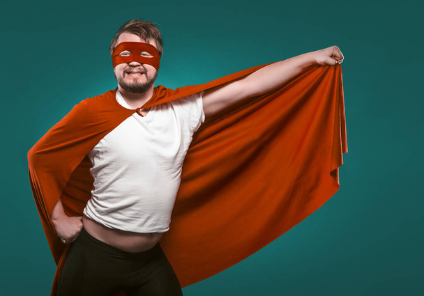 Funny Super Hero Man Ready To Fly Save World. Smiling Man In Mask And Red Superhero Costume Holding His Cloak In His Hand Posing On Biscay Green Background - Photo, Image