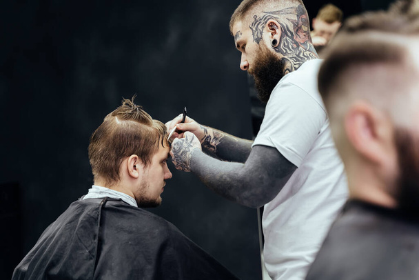 Peluquero tatuado profesional usando afeitadora degolladora cortar el cabello. Hombre atractivo está consiguiendo un corte de pelo moderno en la peluquería. De cerca.
. - Foto, Imagen