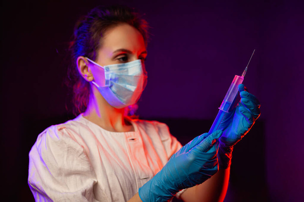 Orvosi munkás orvosi maszkban, fecskendővel a kezében. fiatal orvos lány egy fecskendővel egy sötét szobában, piros és kék fénnyel. orvosi dolgozó a coronavrius pandémia idején - Fotó, kép