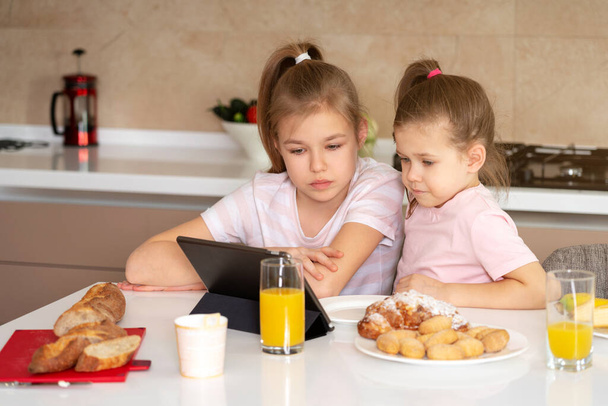 Two sisters having breakfast and watching cartoons on tablet together, happy family concept - Photo, Image