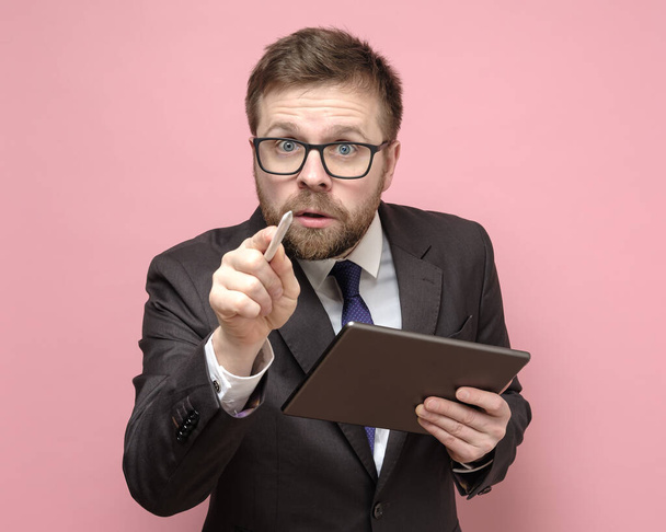 Homme d'affaires caucasien dans un costume avec une tablette regarde curieusement et montre avec un stylet à l'appareil photo
. - Photo, image