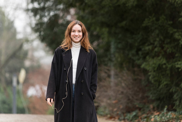 Молодая женщина в длинном черном шерстяном пальто улыбается перед камерой в общественном парке
. - Фото, изображение