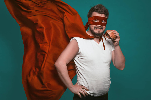 Hungry super hero man eating a hamburger, super hero or antihero in a red suit with a flying cloak eats fast food looking at the camera. Cut out on a green background - 写真・画像
