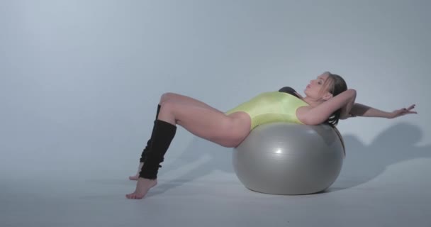 Beautiful girl in a yellow sports suit and black leggings lies on a ball, she is engaged in gymnastics on a gray rubber ball, swimsuit, beautiful skin, figure, barefoot, long hair in the tail - 映像、動画