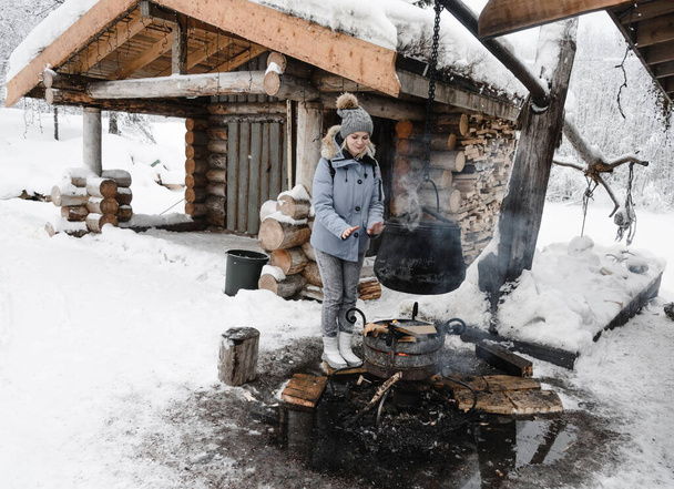 una chica con una chaqueta azul se calienta las manos sobre una hoguera donde se cocina un caldero contra una choza de madera. Karelia invierno
 - Foto, imagen