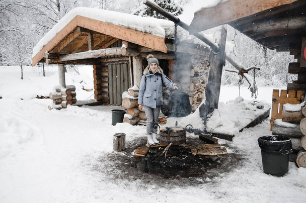 een meisje in een blauw jasje warmt haar handen op over een kampvuur waar een ketel tegen een houten hut wordt gekookt. Karelië winter - Foto, afbeelding